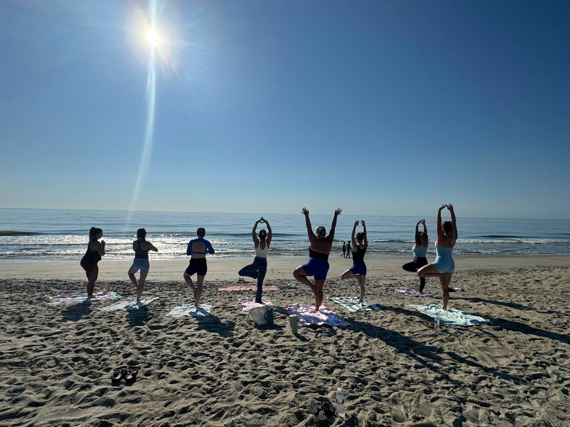 A group practicing tree pose on the sand facing the ocean during a morning beach yoga class